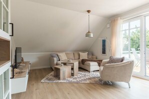 Ein modernes Wohnzimmer mit einer Sofagarnitur, einem Couchtisch und einem Sessel. Ein Fernseher steht auf einem Schrank, ein Fenster mit Vorhängen und eine Deckenleuchte sind ebenfalls zu sehen.
