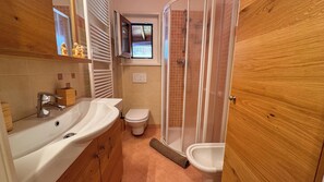 Ein Badezimmer mit Waschbecken, Toilette und Dusche. Eine Holztür und ein Fenster befinden sich über der Toilette.