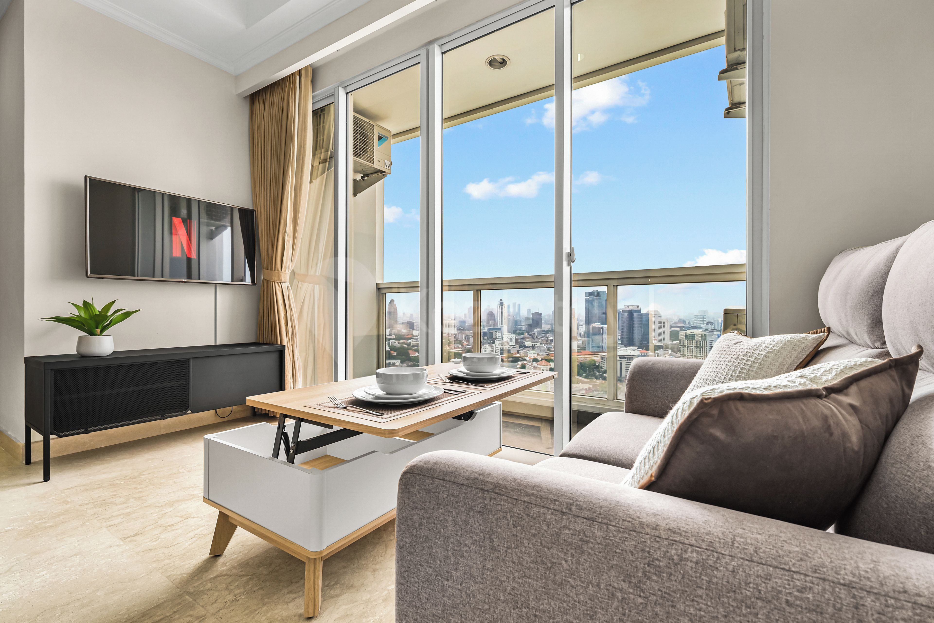 A modern living room with a city view, a flat-screen TV, a dining table with a bowl and spoon, a sofa, and a potted plant.