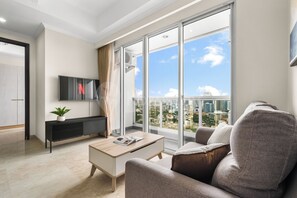 A modern living room with a large window, a flat-screen TV, a sofa, a coffee table, and a plant. - Saira by Kozystay - Menteng (Central Jakarta)
