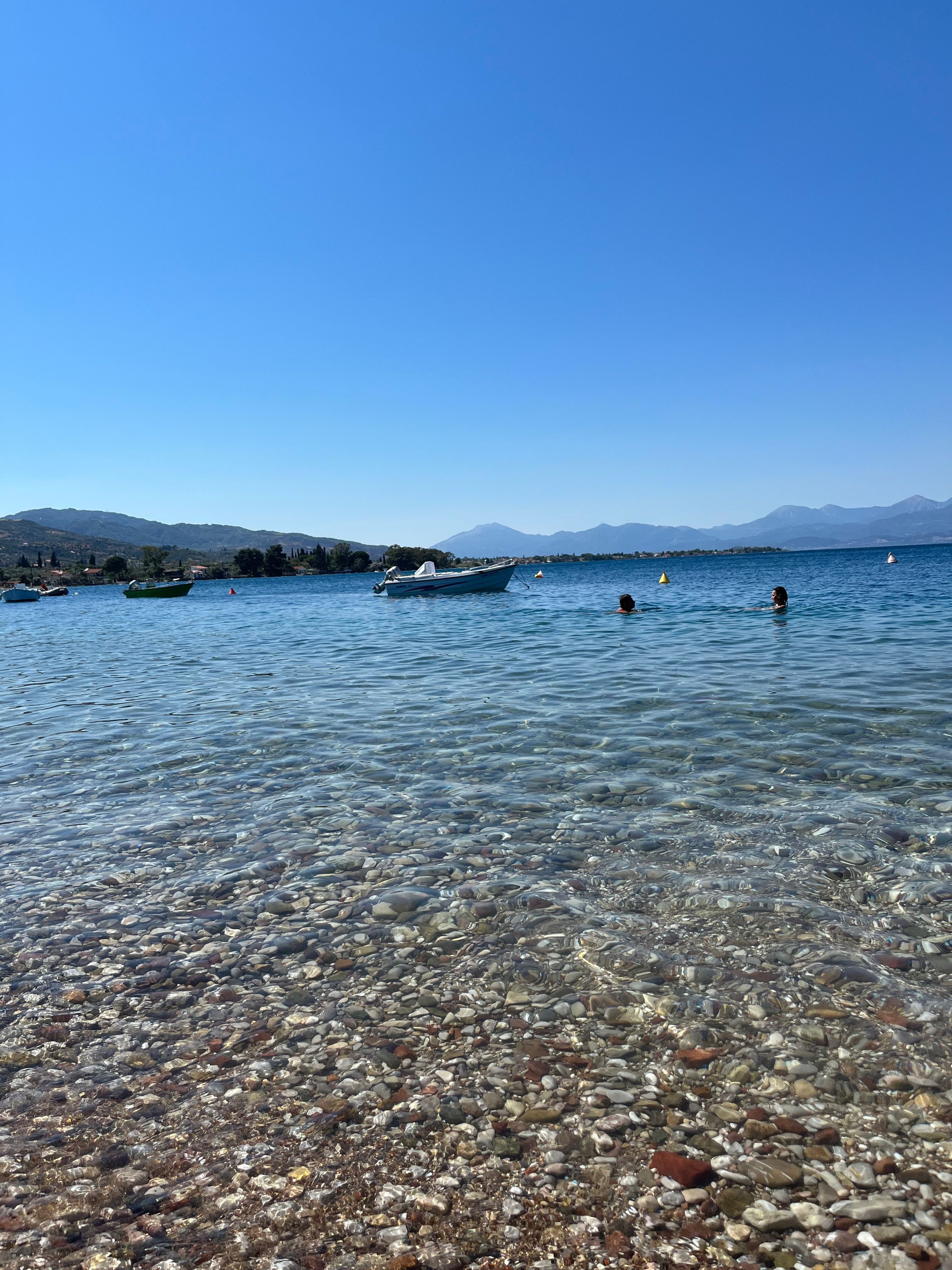 Ein klarer See mit sichtbaren Kieselsteinen und Booten, umgeben von Bergen.