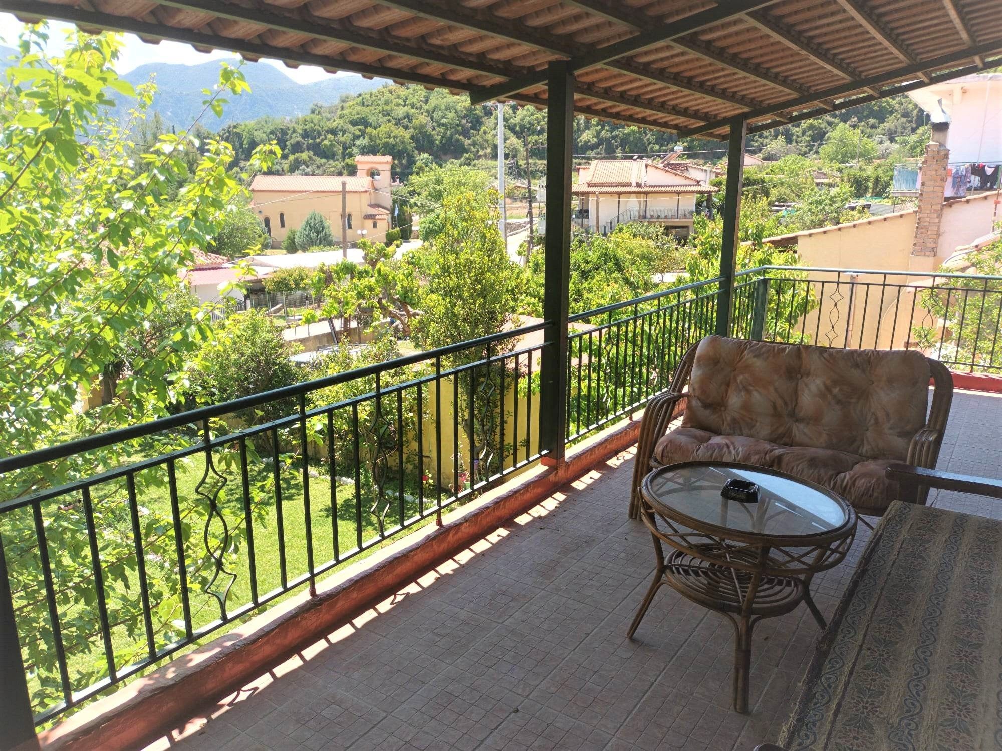 A balcony with a wicker sofa and a glass table, overlooking a green landscape with trees and buildings.