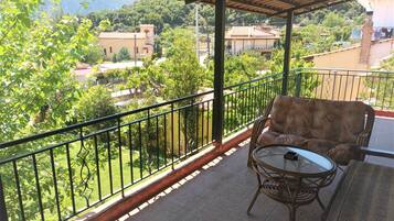 A balcony with a wicker sofa and a glass table, overlooking a green landscape with trees and buildings.