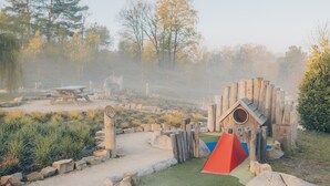 En lekplats med en lekställning i trä, en röd klättervägg och ett picknickbord, belägen i en parkmiljö.