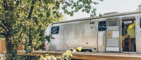 A stationary RV with a door and windows, parked on a wooden deck surrounded by trees.