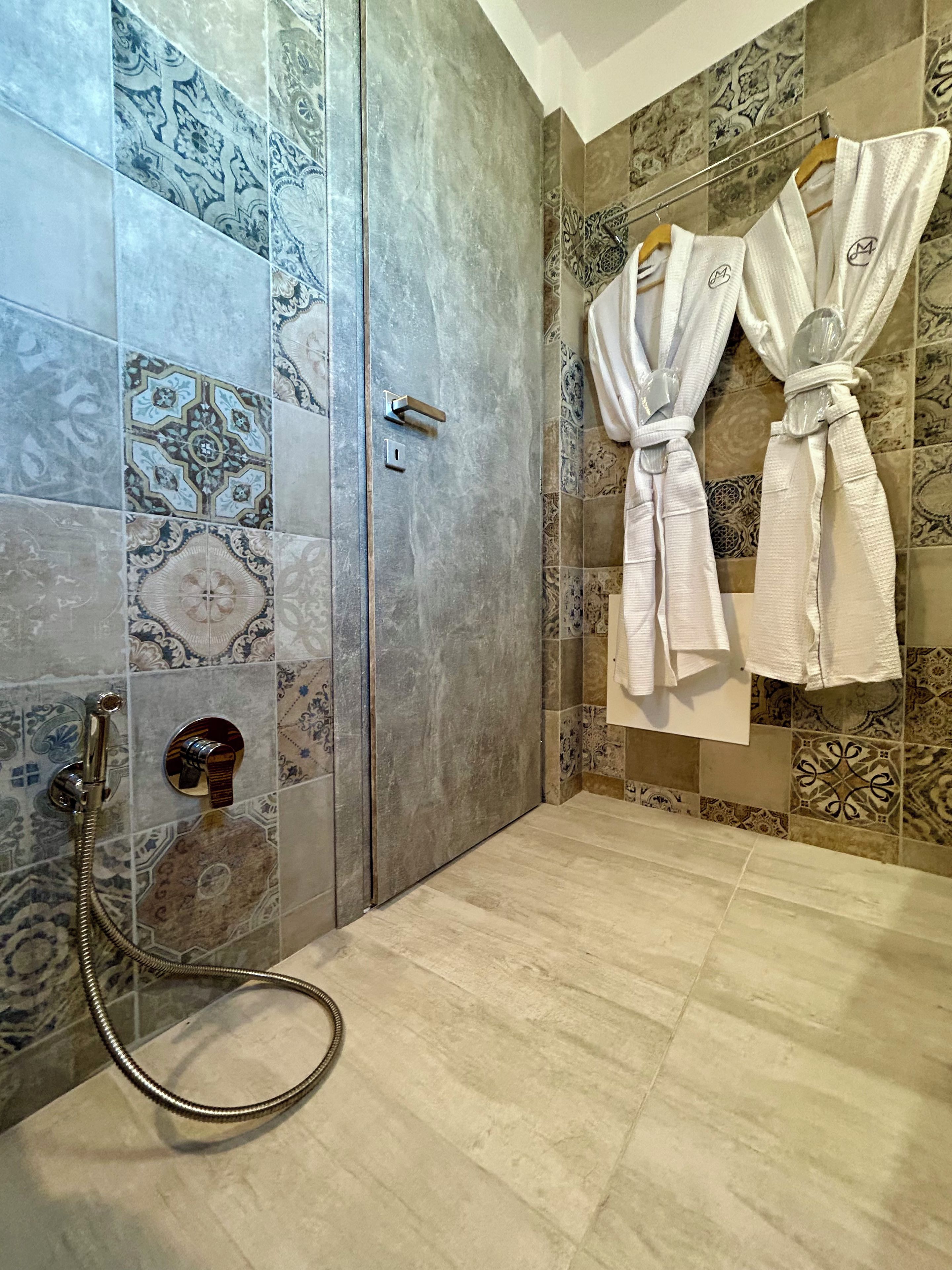 A bathroom with patterned tiles, a shower with a handheld showerhead, and two white robes hanging on a rack.