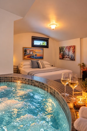 A hotel room with a bed, a painting, a television, and a hot tub with glasses of wine.
