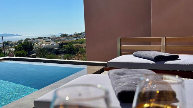 A poolside area with two glasses of wine, a bed, and a view of the sea and distant buildings. - Where luxury meets the magic of Santorini, with a private pool and private spa! (Kamari)