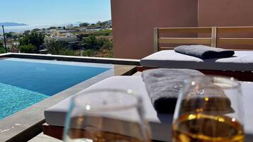 A poolside area with two glasses of wine, a bed, and a view of the sea and distant buildings.