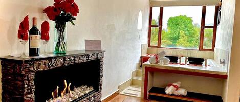 A room with a fireplace, a window with a view, a wooden floor, and a small table with a vase of flowers.