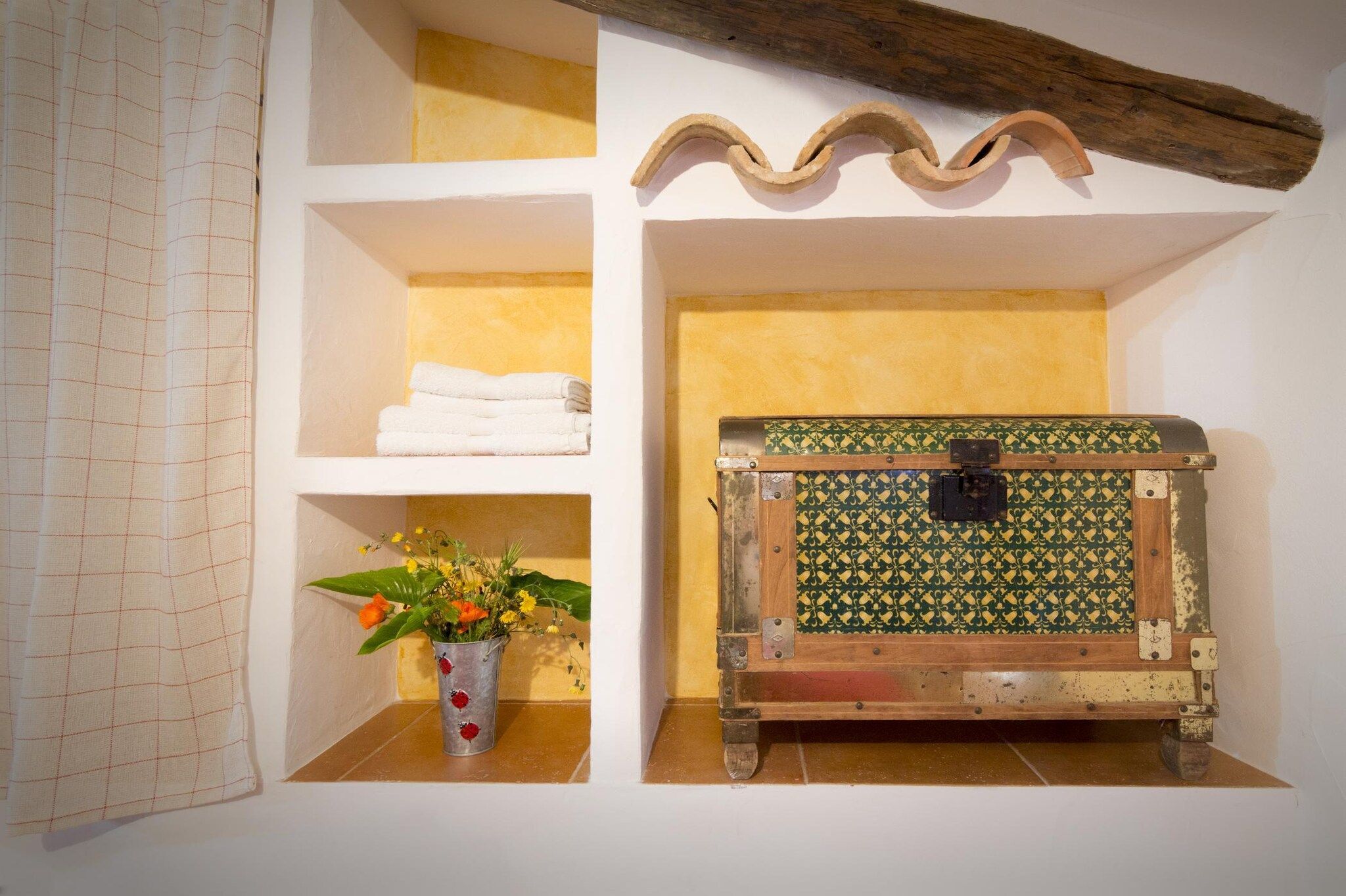 A bathroom with a white tiled wall, a wooden beam, a shelf with folded towels, a metal bucket with flowers, and a patterned trunk.