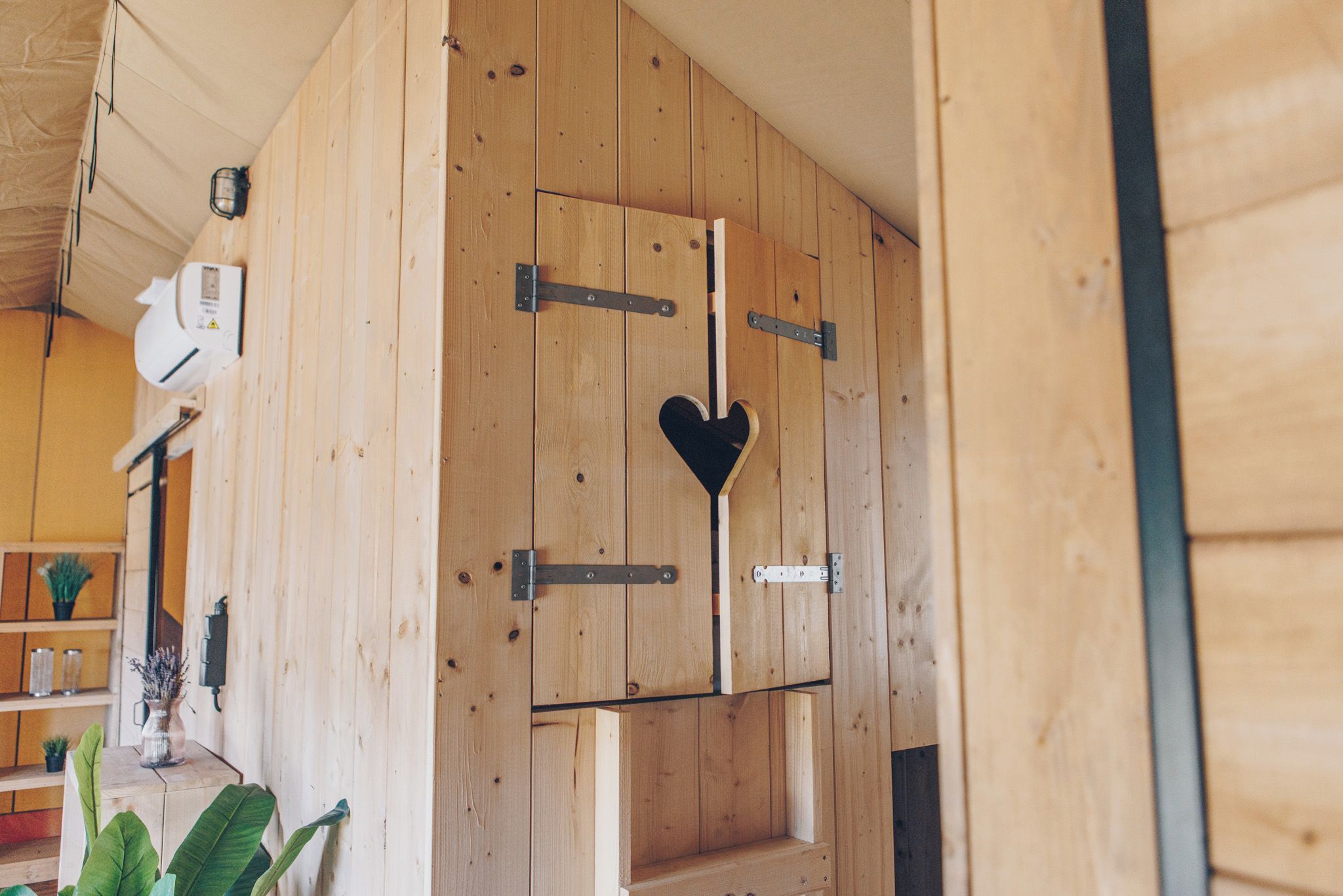 Een houten interieur met een centrale deur, voorzien van een hartvormige uitsparing, metalen scharnieren en een klein plankje.