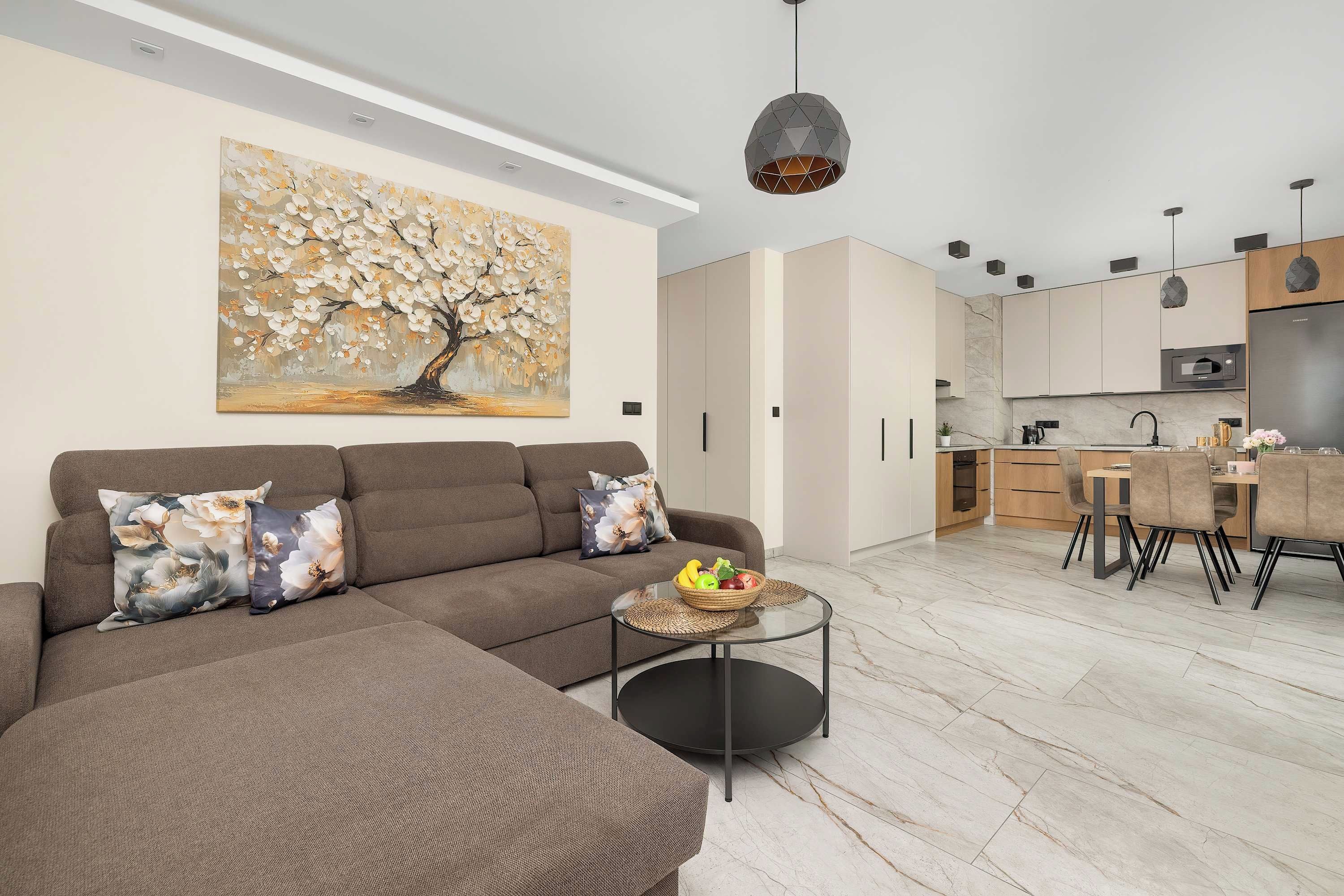 A modern living room with a sectional sofa, a dining area with a table and chairs, a kitchen with a microwave, and a large floral painting on the wall.