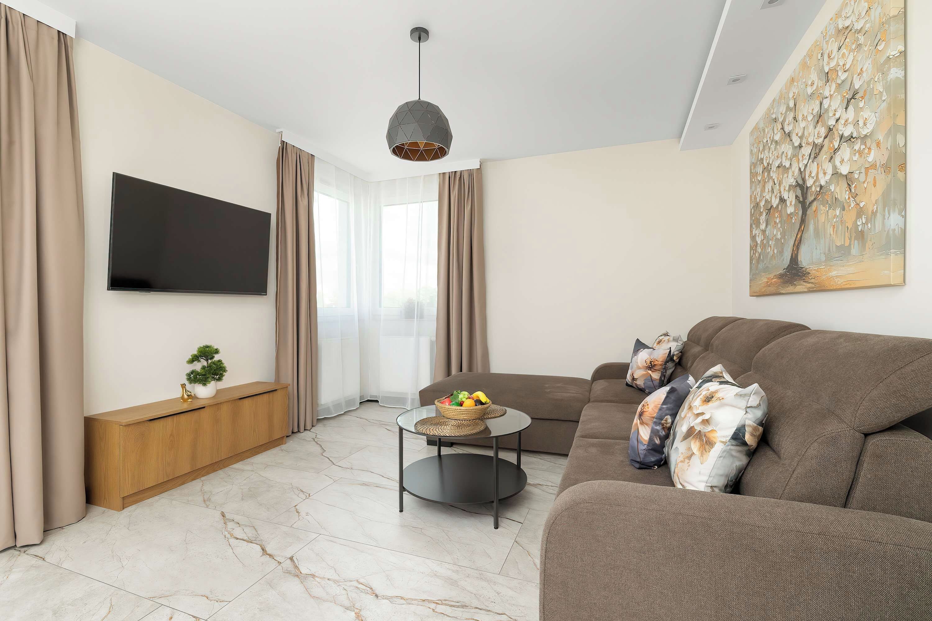A modern living room with a flat-screen TV mounted on the wall, a brown sofa with decorative pillows, a small round coffee table with a fruit bowl, and a painting on the wall.