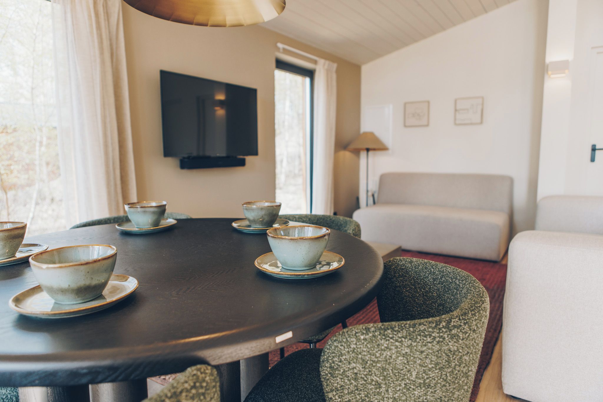 A round table with six bowls and saucers, a sofa, a TV mounted on the wall, and a window with curtains.