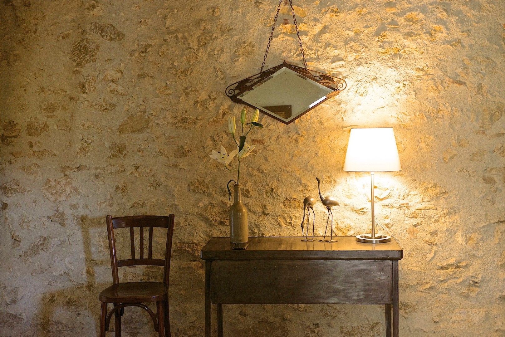Una habitación rústica con una silla de madera, una mesa con lámpara, un florero con flores y un espejo colgado.