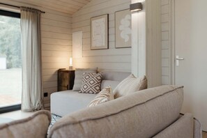A bedroom with a bed, pillows, a bedside table, and a lamp. There are framed pictures on the wall and a window with curtains.