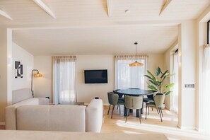 A living room with a sofa, a dining table with chairs, a TV mounted on the wall, and a potted plant.