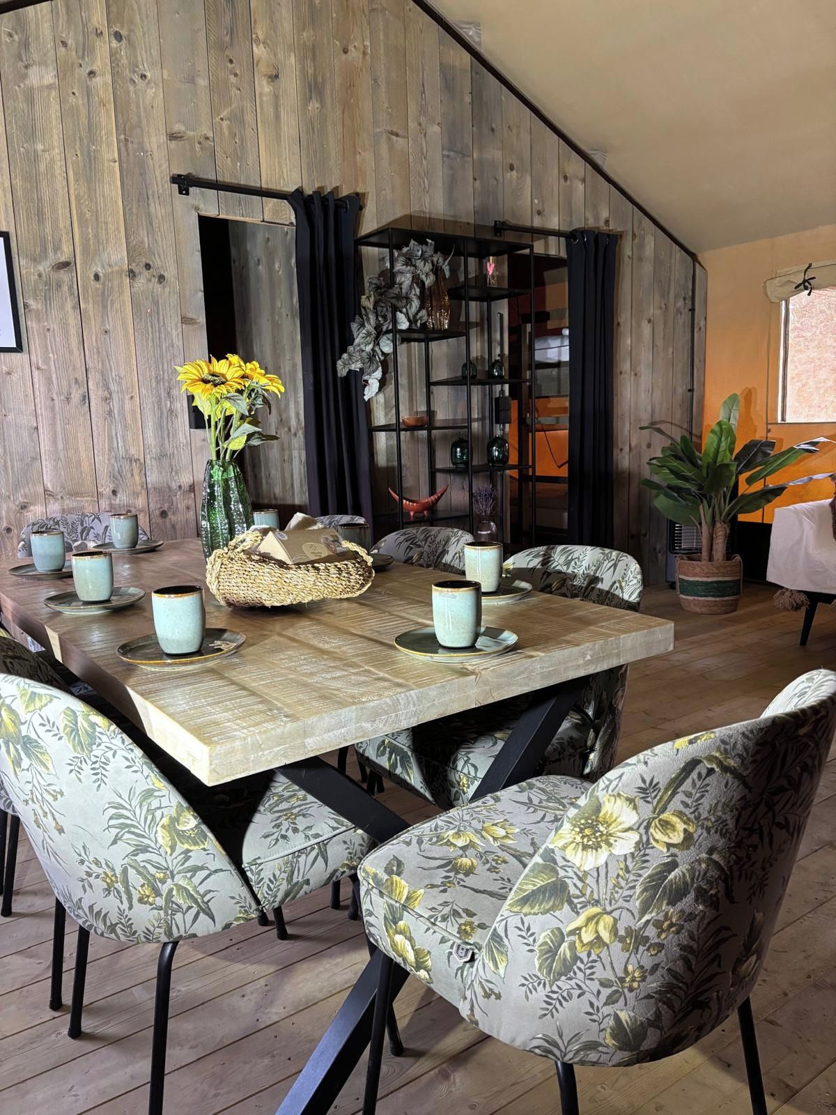 Ein Essbereich mit einer Holzwand, einem schwarzen Metallregal und stuhlförmigen Sitzgelegenheiten mit floralem Muster.