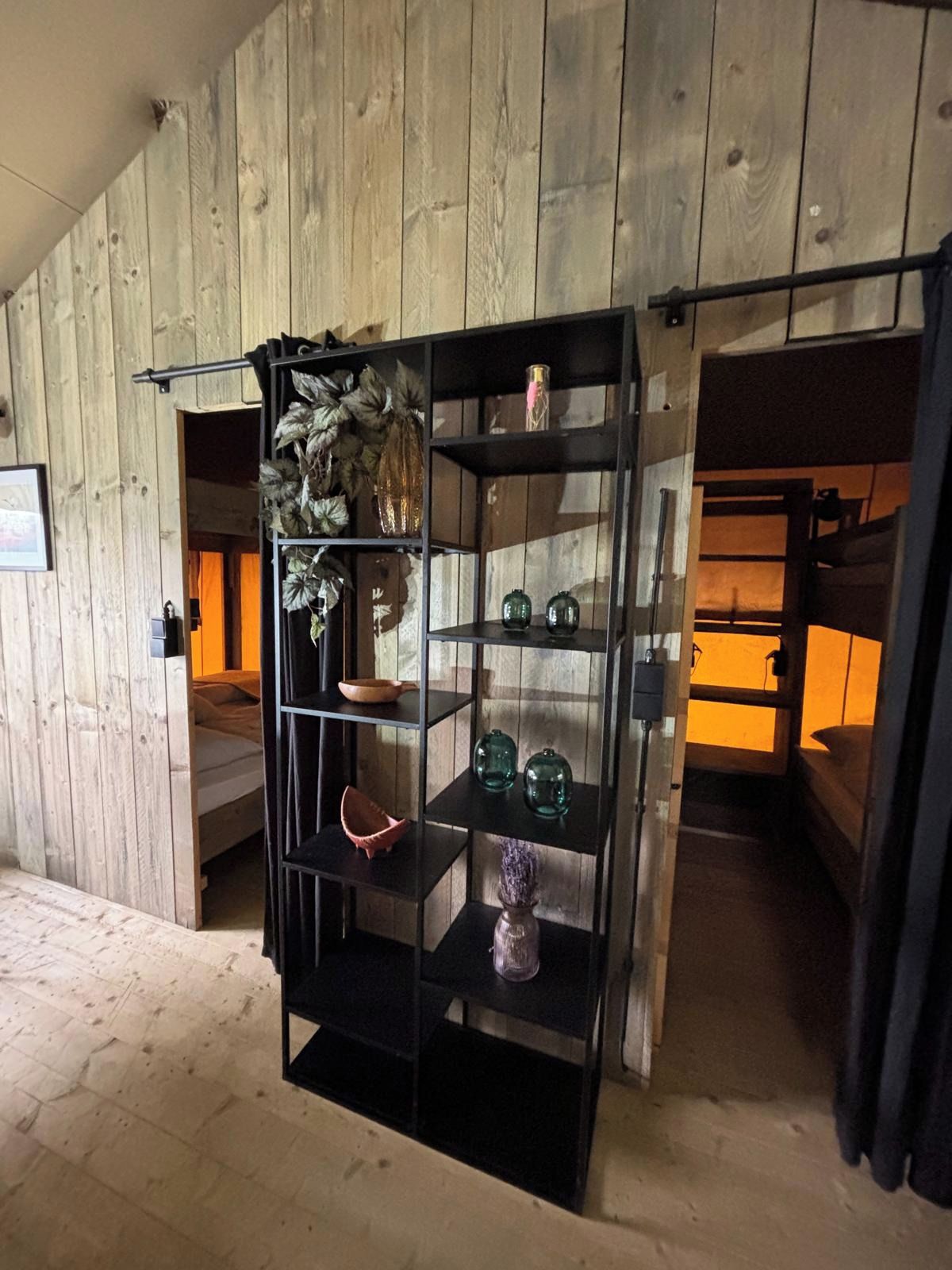 Une pièce avec un mur en bois, une étagère noire ornée d’objets décoratifs et un lit avec une tête de lit.