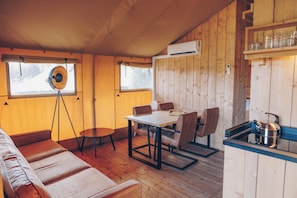 Interior de una cabaña de madera con una mesa de comedor, sillas, un sofá, una mesita redonda, un equipo de aire acondicionado y una tetera sobre una encimera.