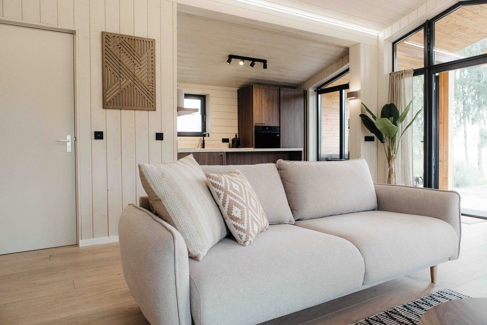 Un salon moderne avec un canapé, un panneau décoratif au mur et une vue sur la cuisine et l’extérieur.