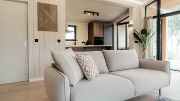 Un salon moderne avec un canapé, un panneau décoratif au mur et une vue sur la cuisine et l’extérieur.