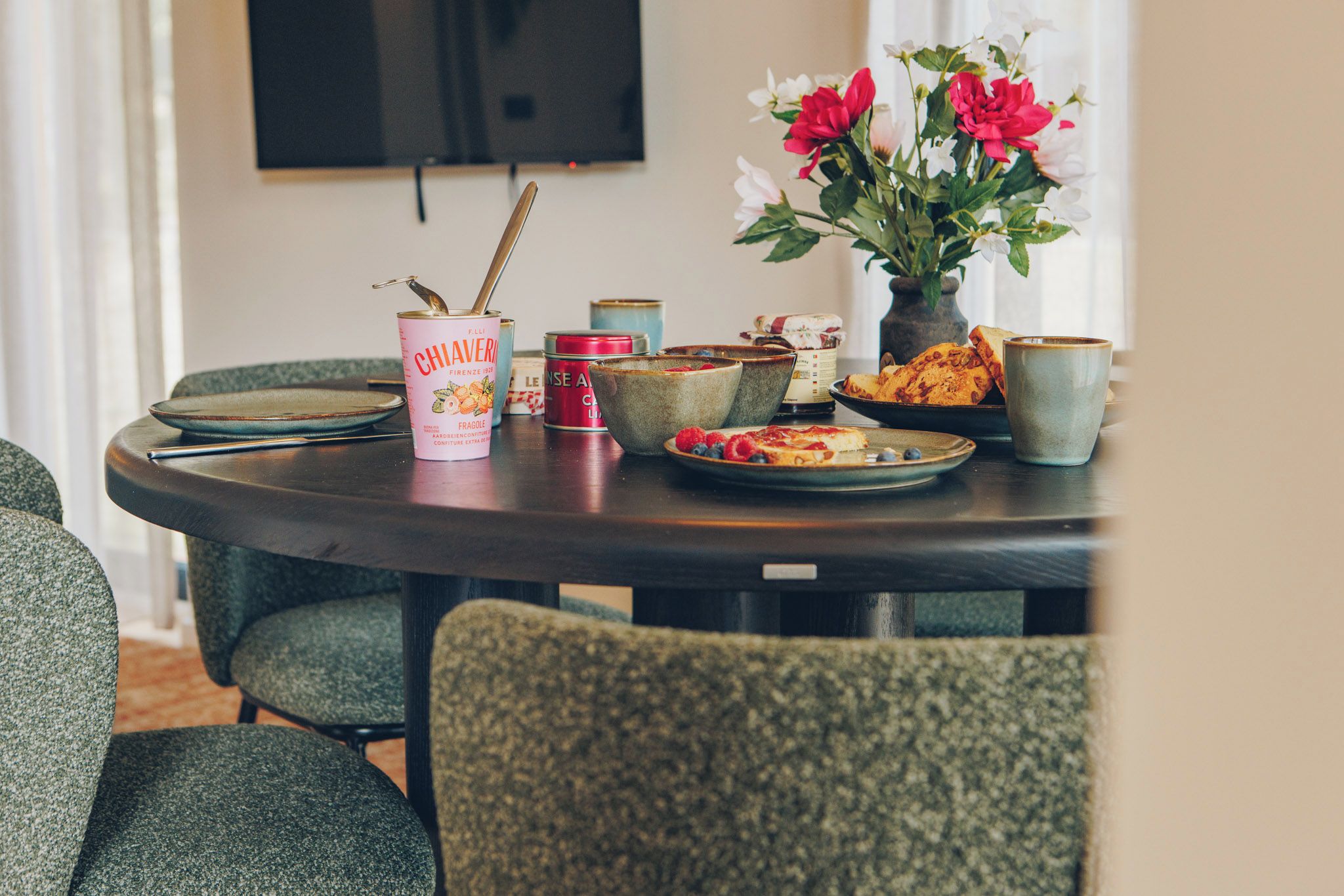 A dining table with a variety of food items, including a can of Chiaverini, a plate with bread and berries, and a vase with flowers.