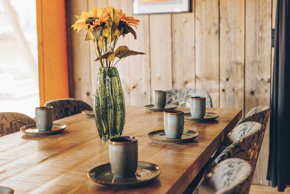 A wooden dining table with a glass vase containing flowers, surrounded by chairs and plates with cups.