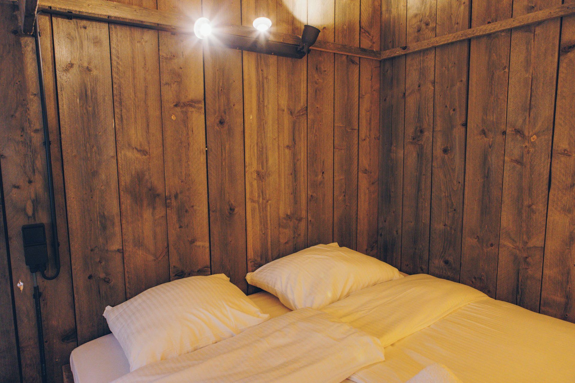 A wooden room with a bed, two wall-mounted lights, and a small shelf.