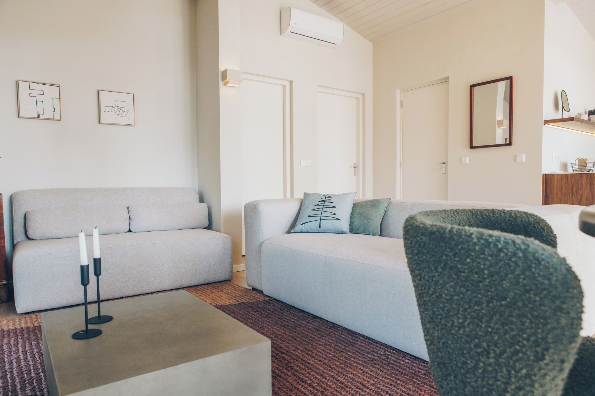 Ein modernes Wohnzimmer mit einem grauen Sofa, einem Couchtisch und einem grünen Sessel. Zwei Türen und eine Klimaanlage sind an der Wand angebracht.