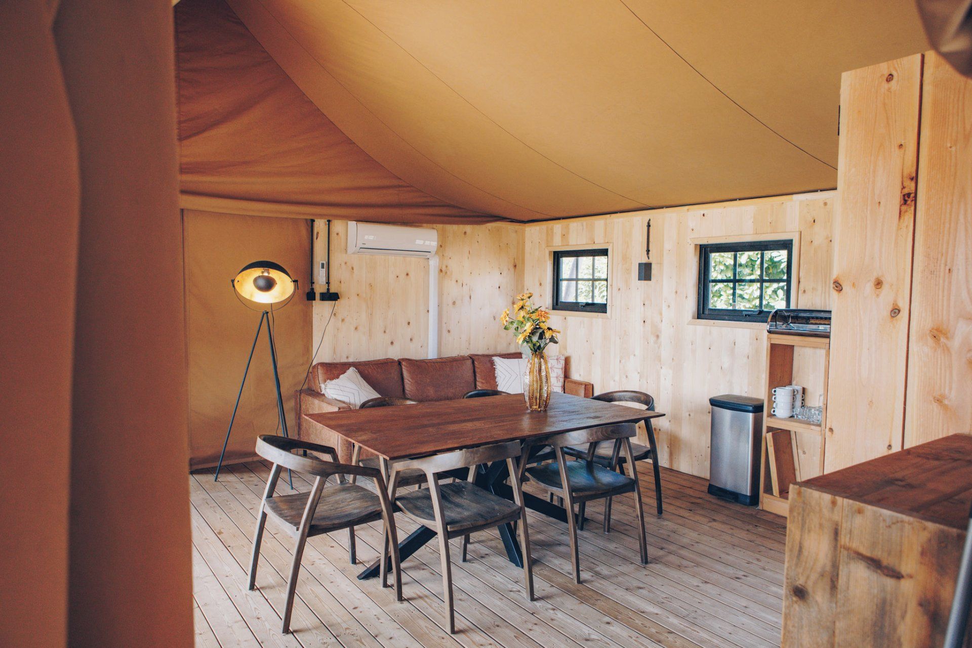 Ein gemütliches Holzhaus-Interieur mit Esstisch, Stühlen, einer Couch, einer Stehlampe und einer kleinen Küchenzeile.