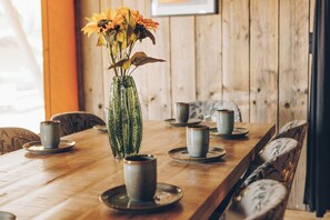 A wooden dining table with a glass vase containing flowers, surrounded by chairs and plates with cups.