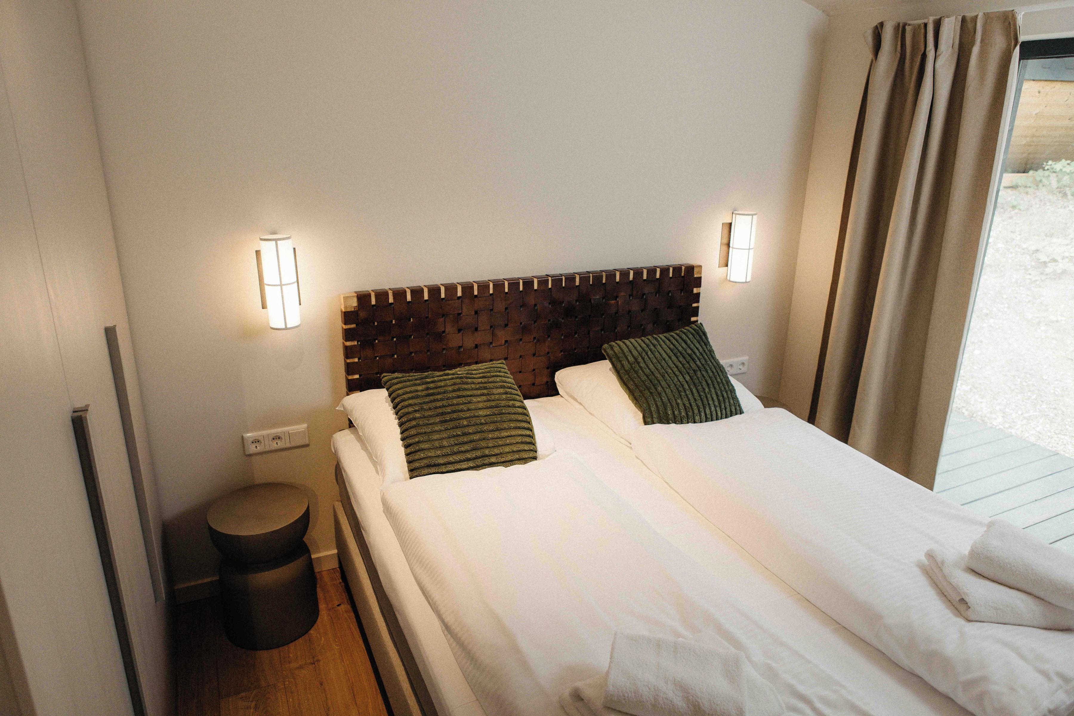 A neatly made bed with white linens, two pillows, a round bedside table, and two wall-mounted lights.