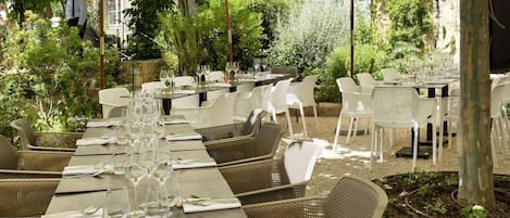An outdoor dining area with tables and chairs, surrounded by greenery and a stone wall.