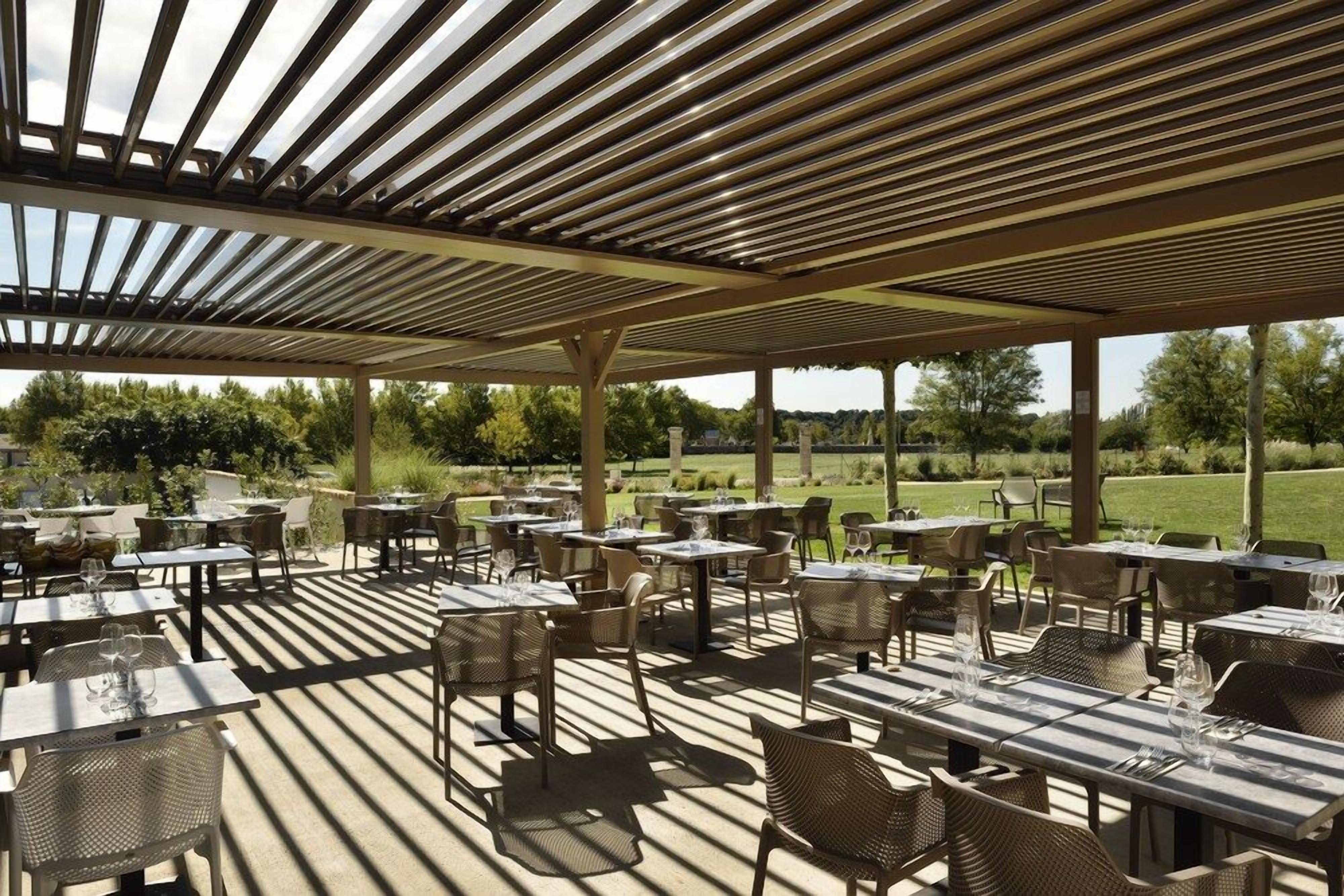 An outdoor patio with tables and chairs, a pergola overhead, and a view of a green landscape.