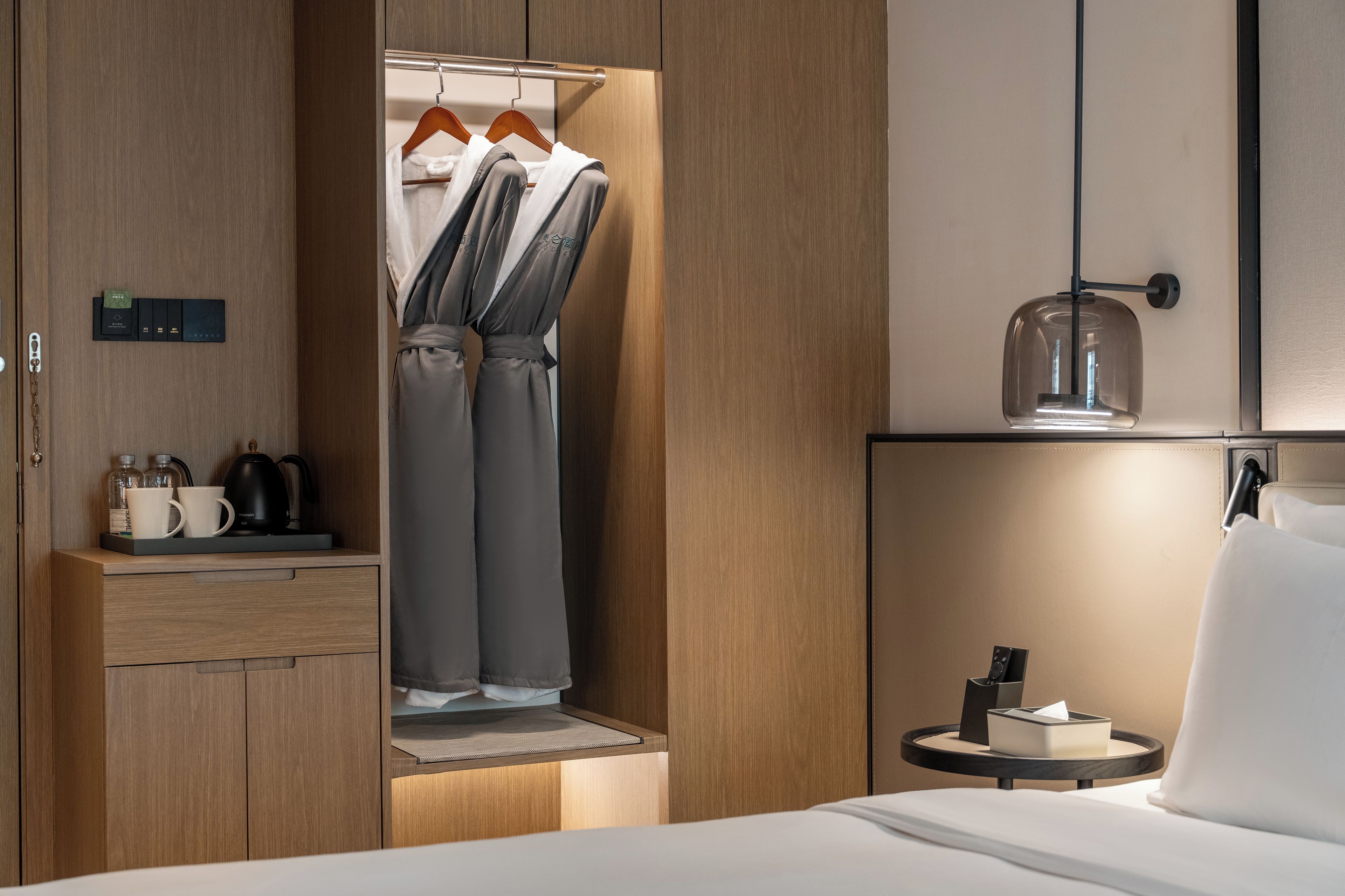 Habitación de hotel con un armario de madera, una cama, una mesita de noche con lámpara y una pequeña mesa redonda con una caja de pañuelos.