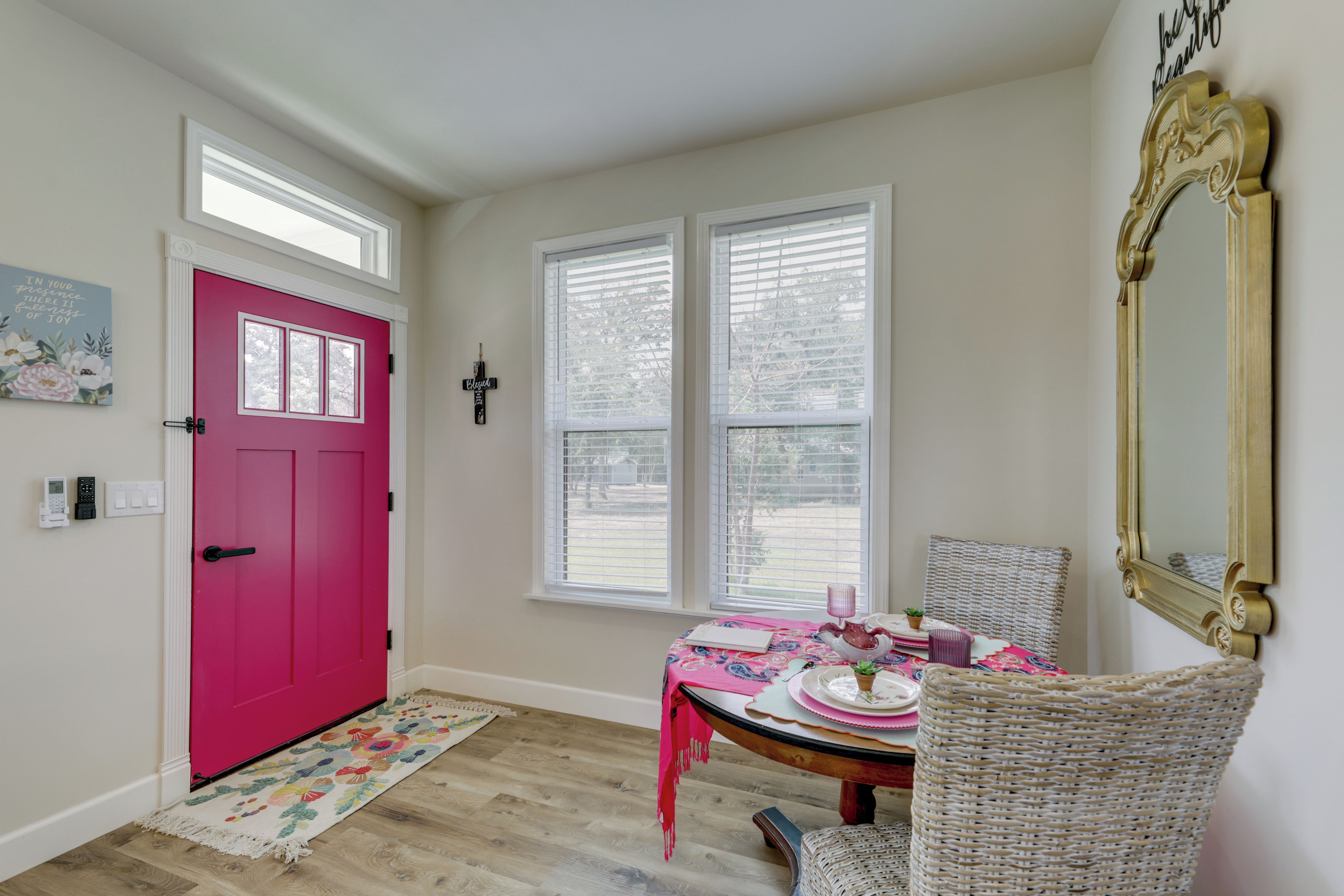 Una habitación con una puerta rosa, dos ventanas con persianas, una mesa con mantel rosa, una silla de mimbre y un espejo en la pared.