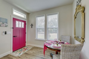 Ein Zimmer mit einer rosafarbenen Tür, zwei jalousieförmigen Fenstern, einem Tisch mit einer rosa Tischdecke, einem Korbstuhl und einem Spiegel an der Wand.