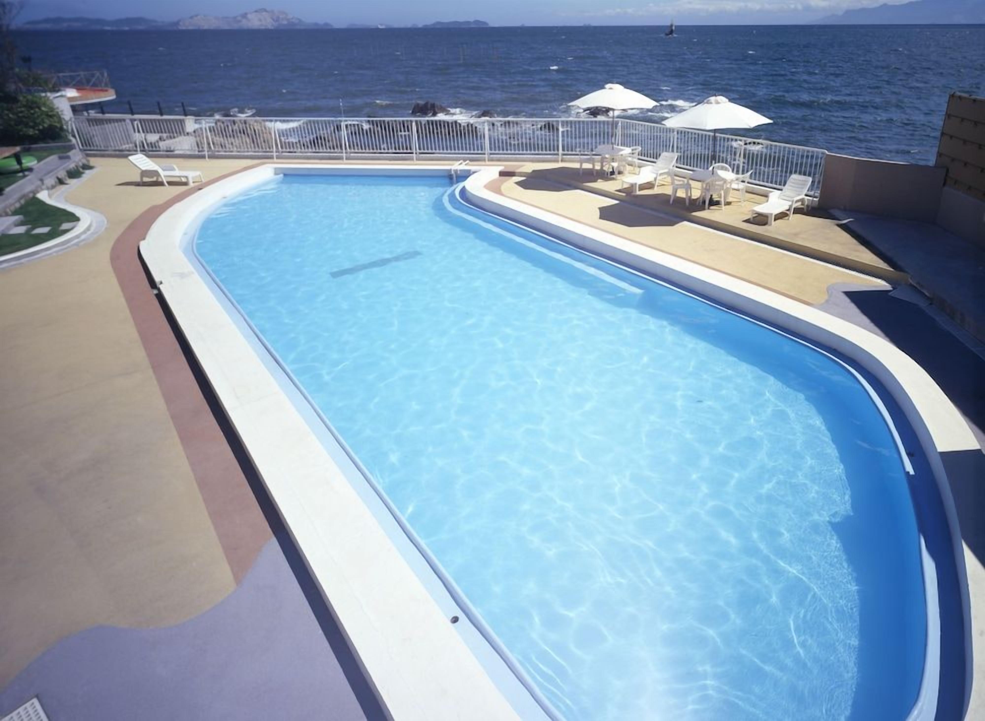 Una piscina con borde curvo, rodeada de sillas de descanso y sombrillas, con vista al mar y una isla distante.