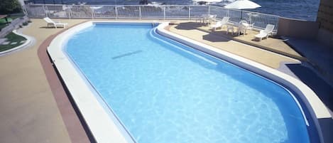 Una piscina con borde curvo, rodeada de sillas de descanso y sombrillas, con vista al mar y una isla distante.