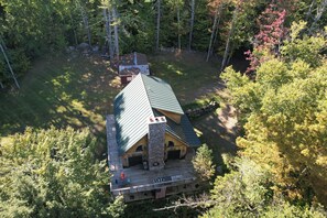 A lodge with a stone chimney, a porch with outdoor seating, and a view of the surrounding forest.
