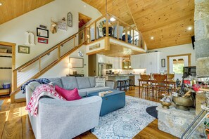 A spacious living area with a high ceiling, a staircase leading to an upper level, a kitchen with modern appliances, and a dining area with wooden chairs.