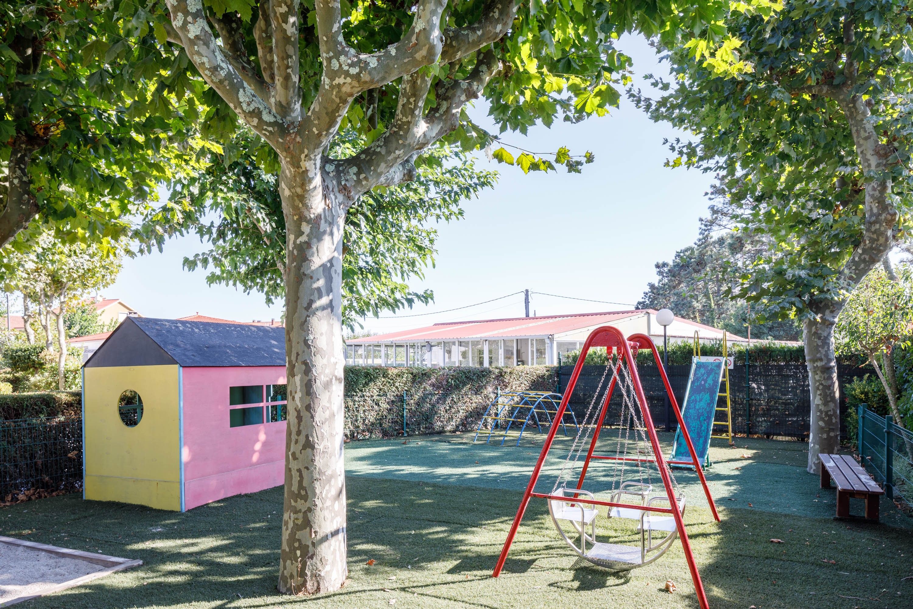 Un parque infantil con columpios y un tobogán, rodeado de árboles y un edificio pequeño.