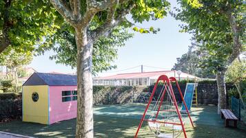 Un parque infantil con columpios y un tobogán, rodeado de árboles y un edificio pequeño.