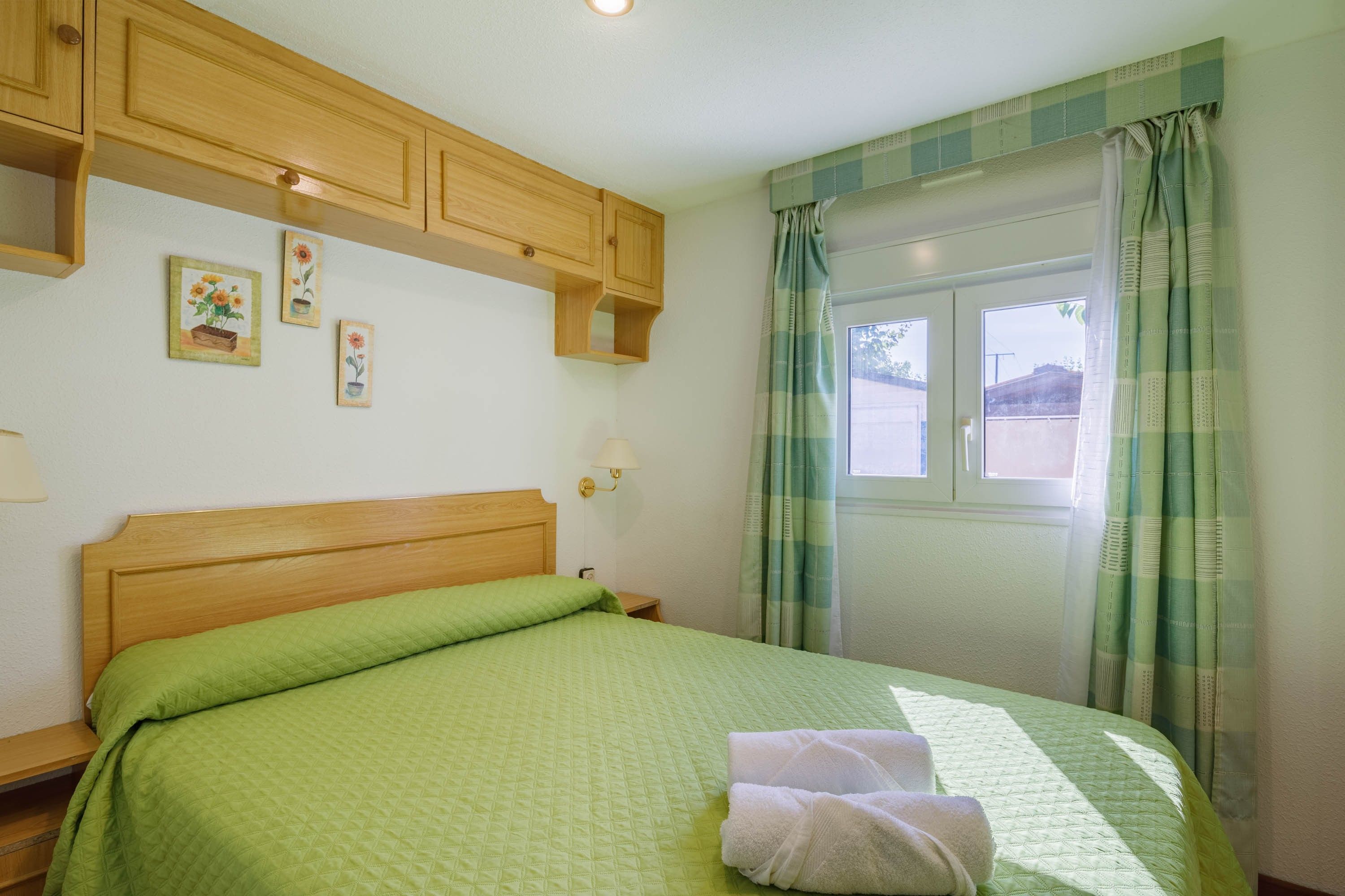 A bedroom with a wooden headboard, a green quilted bedspread, a nightstand with a lamp, and a window with curtains.