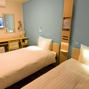 Une chambre d’hôtel avec deux lits simples, un bureau avec une chaise, un miroir et une étagère fixée au mur avec un livre.