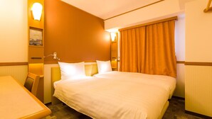 Quarto de hotel com uma cama grande, uma mesa de trabalho e uma cadeira. Há uma janela com cortinas, uma luminária na parede e um pequeno porta-retrato.