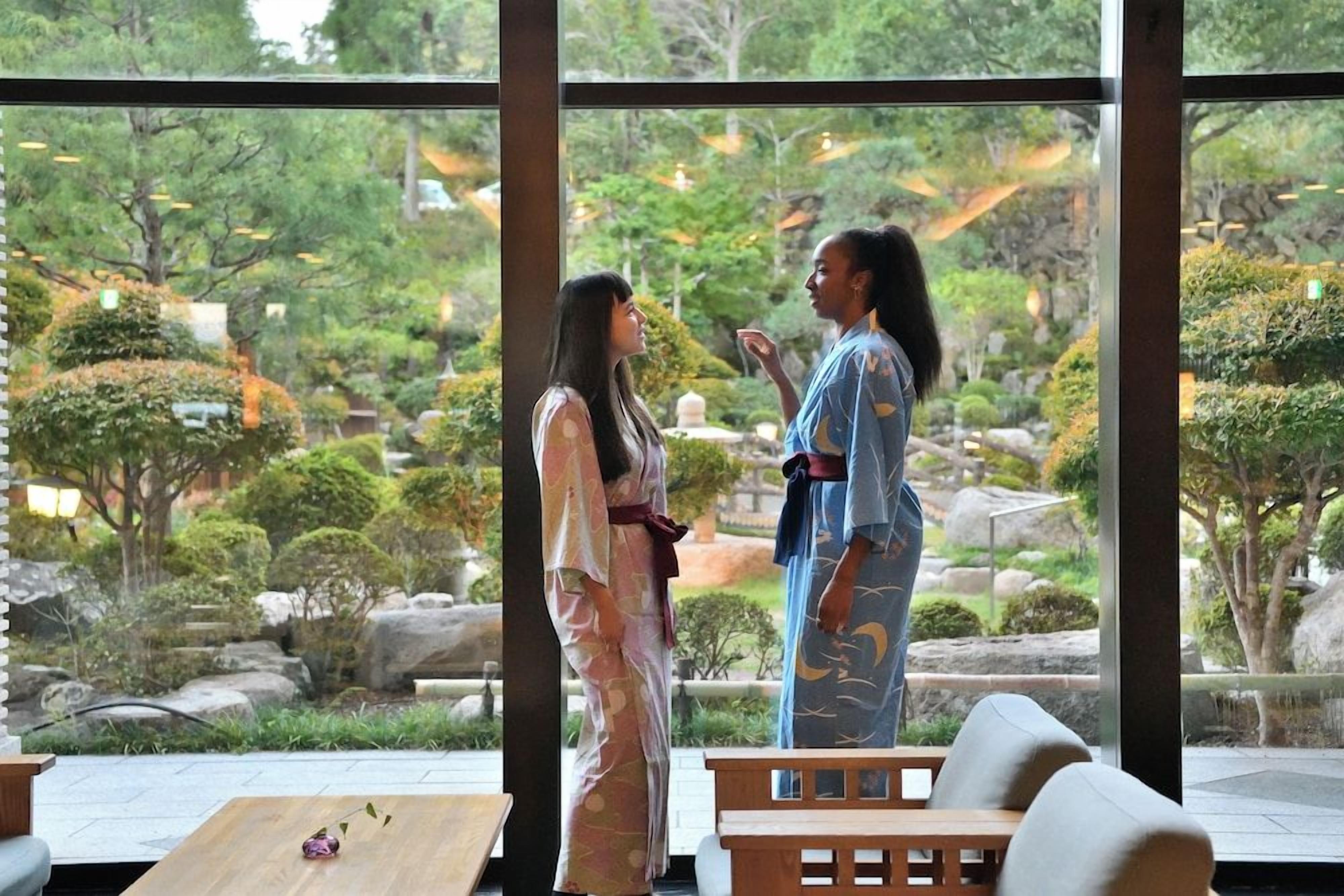 Two individuals in traditional attire standing in a room with a view of a landscaped garden through large windows.