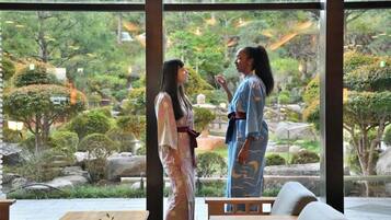 Two individuals in traditional attire standing in a room with a view of a landscaped garden through large windows.